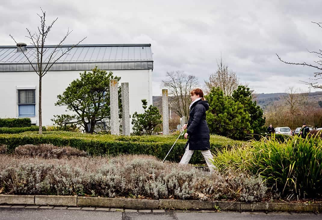 A woman (Sandra Pilz) who is blind using her white cane to walk on the green grounds of the BFW campus, with rolling hills in the background.