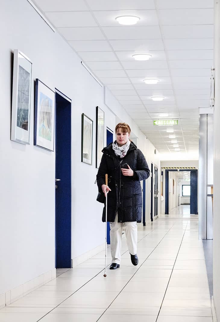 A woman (Sandra Pilz) who is blind using her white cane to guide her through the corridor of BFW Würzburg
